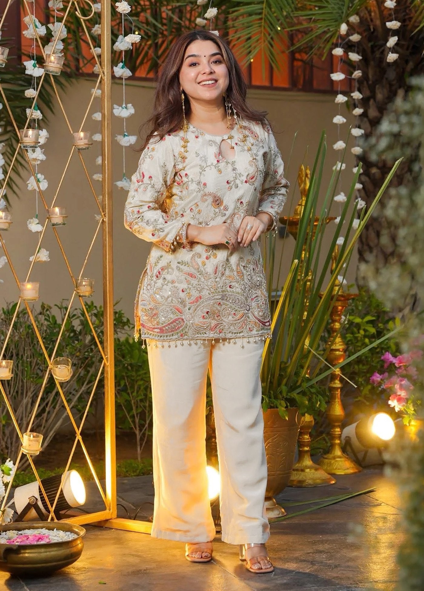 Woman in a floral top and white pants standing in an decorated outdoor setting with plants and lights.